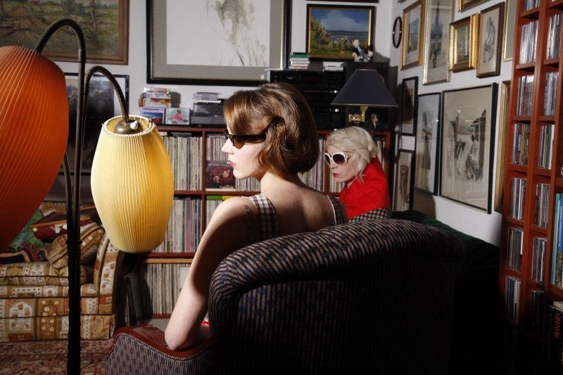 Model mit Kurzhaarfrisur und Sonnenbrille in einem Wohnzimmer voller Bilder und Lampen, daneben eine ältere Dame