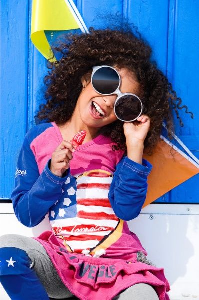 Mädchen mit Locken und großer Sonnenbrille lacht vor einer blauen Strandhütte