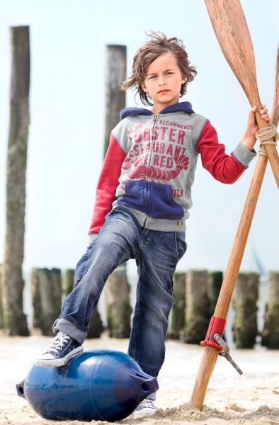 Junge mit Paddel im Kapuzenpullover zwischen Holzpfählen am Strand
